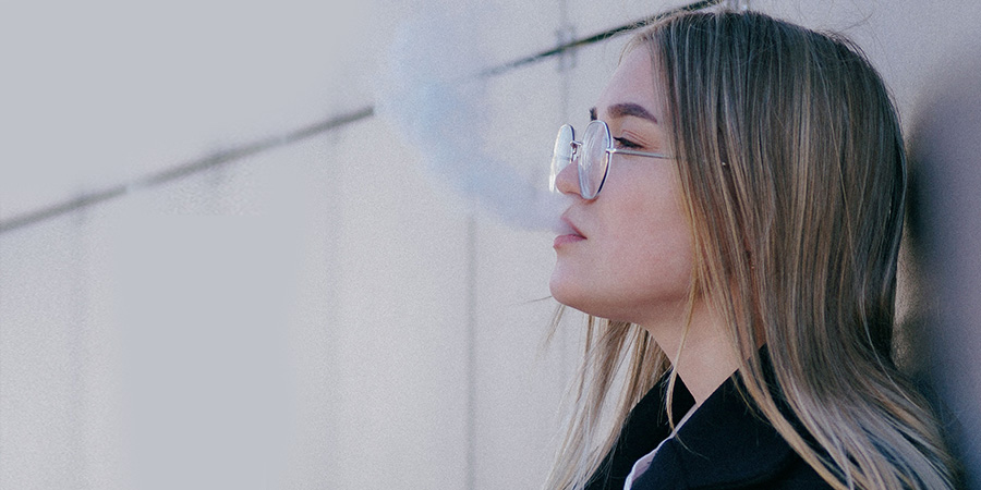 woman with long blonde straight hair wearing glasses and a black coat leaning on a wall, vaping and exhaling smoke
